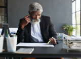 mature business man with beard seated at desk, pen in hand, studying information on paper